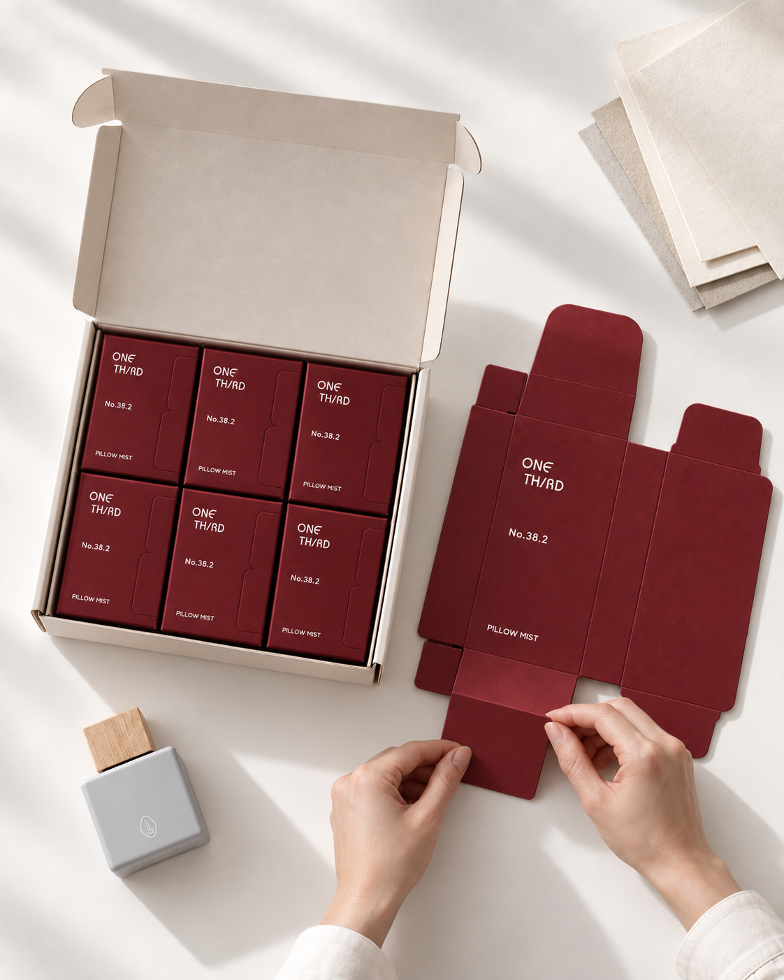 A styled flat-lay of a product set showing several small red boxes inside an open beige cardboard box, alongside matching red packaging flaps and a small gray stamp-like tool. Two hands are visible assembling one of the red packaging pieces on a white surface with soft shadows.